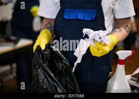 Young adult Caucasian man wearing yellow gloves cleaning office workspace, holding crumpled paper and napkin while disposing trash into black garbage bag, spray bottle visible nearby Stock Photo