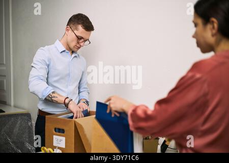 Young adult Caucasian man sorting documents into cardboard box while young adult woman assisting in office cleaning process, both focused on organizing workspace during cleanup Stock Photo