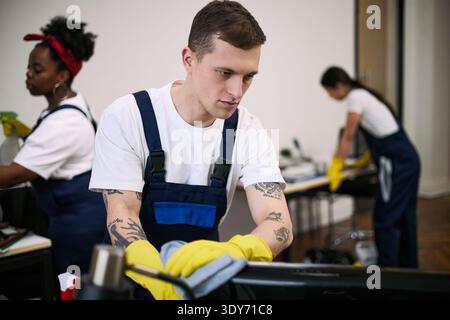 Caucasian young adult man cleaning office desk with yellow gloves while Black young adult woman and Asian young adult woman, working in background performing cleaning tasks Stock Photo