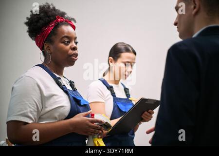 Black young adult woman holding cleaning supplies and talking to Caucasian man while Asian young adult woman writing on clipboard in office setting, both women wearing work uniforms Stock Photo