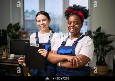 Two young adult women, one Caucasian and one Black, standing together in office wearing cleaning uniforms, smiling at camera, holding clipboard, representing cleaning staff Stock Photo