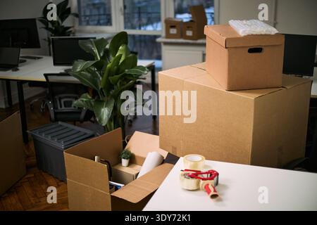 Stacked cardboard boxes and cleaning supplies occupying modern office workspace, packing materials and tape dispenser arranged on desk, indoor plant visible among moving items Stock Photo