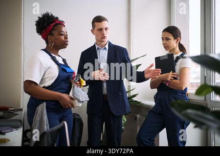 Caucasian young adult man instructing Black young adult woman and Hispanic young adult woman in cleaning uniforms, holding cleaning supplies and clipboard in modern office setting Stock Photo