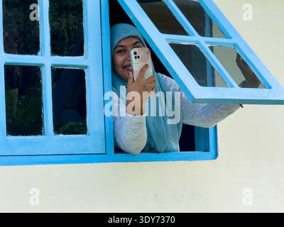 Woman uses cell phone while looking out of a window in Banda Neira, Moluccas, Indonesia, smiling and engaging with someone Stock Photo