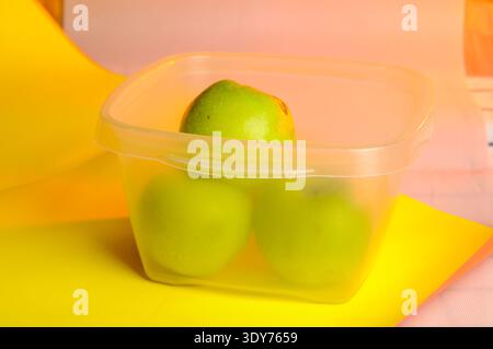 A close up view shows three vibrant green apples inside a transparent square storage box, set against a colorful backdrop Stock Photo