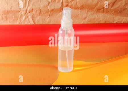 A transparent plastic spray bottle containing clear liquid stands against a backdrop of crumpled brown paper and rolls of red and orange material Stock Photo