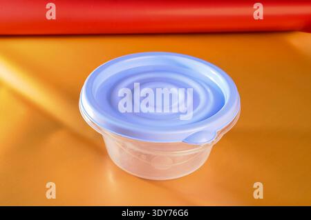 A small, round, transparent food storage container with a textured blue lid sits on a vibrant orange surface Stock Photo