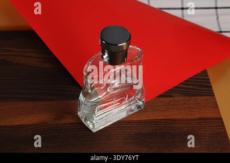 A clear glass perfume bottle with a textured black cap sits on a dark wooden surface, accented by a vibrant red paper background Stock Photo
