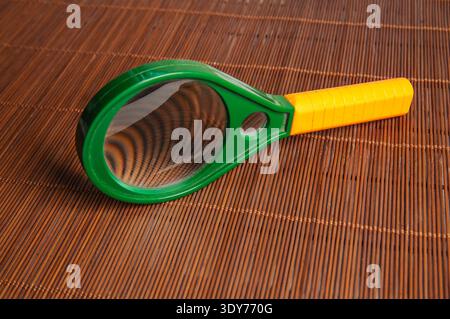 A child's magnifying glass, featuring a vibrant green rim and a textured yellow handle, is positioned on a brown bamboo placemat, highlighting its pla Stock Photo