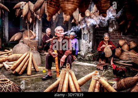 Man crafts bamboo fish traps while smoking in workshop with other artisans in Hanoi, Vietnam Stock Photo