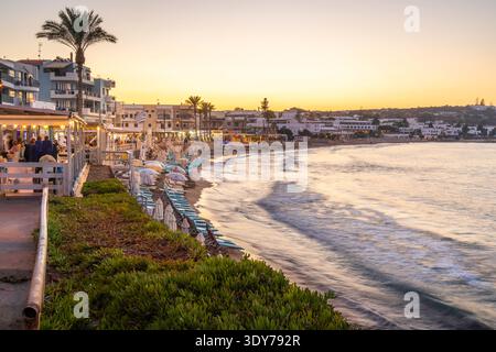 View of Palmowy Zaktek Beach at sunset with buildings and beach chairs in Limenas Chersonisou, Greece Stock Photo