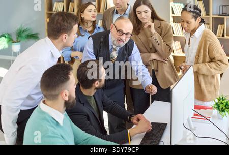 Senior mentor guiding adult students around computer in office training session Stock Photo