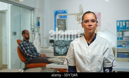 Successful dentist medic having a speech about hygienic teeth, looking at camera, talk trustful in dental clinic while assistant preparing man for stomatological surgery in background. Stock Photo