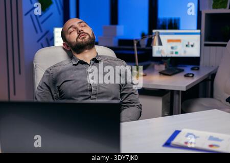 Exhausted hardworking businessman sleeping on chair. Workaholic employee falling asleep because of working late at night alone in the office for important company project. Stock Photo