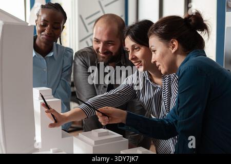 Multi ethnic architectural teamwork analyzing maquette building model construction layout. Diverse professional group of colleagues pointing at project design blueprint plan Stock Photo