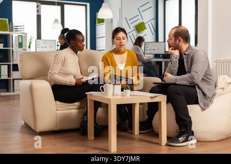 Confident company manager giving working tasks to diverse teammates sitting on couch in start up office. Multiethnic team discussing project ideas at brainstorming meeting using digital devices Stock Photo