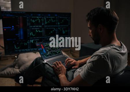 Successful trader engrossed in financial analysis and day trading, using laptop and large external monitor displaying various stock market and Stock Photo