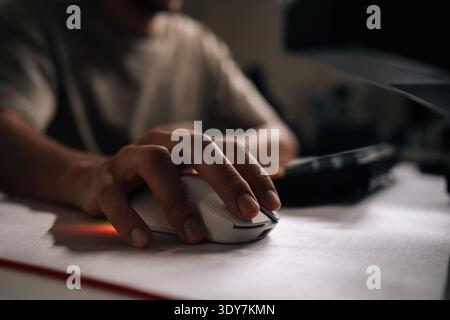 Close-up of male hand using white rgb gaming mouse on white mousepad, with keyboard and monitor in background, suggesting late-night gaming session. Stock Photo