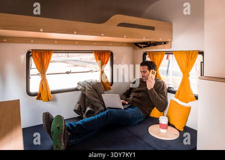 Man talking on phone and typing on laptop inside modern camper van, enjoying nomadic lifestyle and remote work Stock Photo