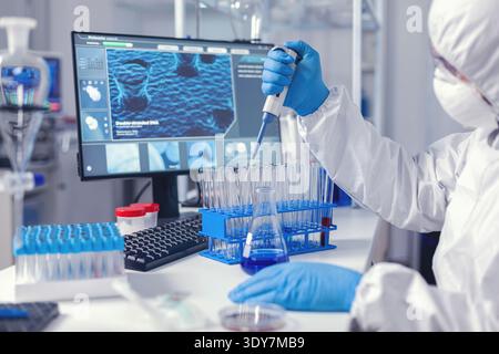 Laborant dropping sample in test tube using micropipette dressed in coverall. Chemist in modern laboratory doing research using dispenser during global epidemic with covid-19. Stock Photo