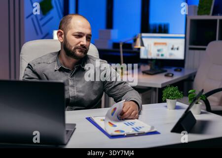 Businessman working late at night in workplace using tablet pc holding hand on clipboard with chart. Bearded entrepreneur doing overhourse at the office in the evening to finish a deadline. Stock Photo