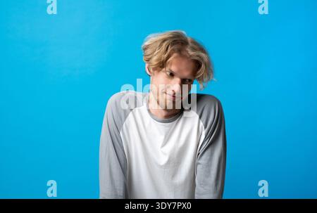 Young man shrugging shoulders, looking away with a quizzical uncertain expression, expressing confusion, doubt, and indecision on a blue background Stock Photo