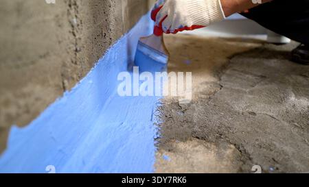 Worker applies blue waterproofing mastic to a concrete basement joint. Close-up on brush and sealant. Textured grey surface contrasts with vibrant blu Stock Photo