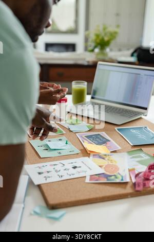 Corkboard is holding printed cards and photos while laptop is displaying spreadsheet near window Stock Photo