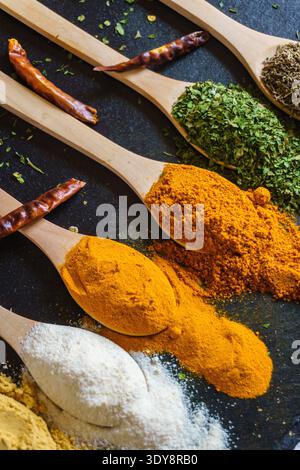 Close-up of wooden spoons filled with colorful spices and dried herbs on dark slate background, including turmeric, curry powder, flour, and dried chi Stock Photo