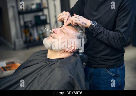 Barber making hair removal procedure in a barbershop for mature man Stock Photo