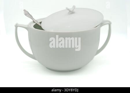 White porcelain soup tureen with lid and two handles, silver spoon resting inside, high-key studio background Stock Photo