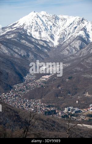 Krasnaya Polyana, Russia - February 21, 2026: Snow covered mountain peak rising above Krasnaya Polyana valley with village buildings and forested Stock Photo