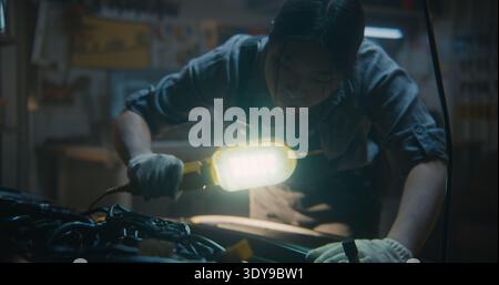 Professional Female Mechanic Conducts Thorough Visual Inspection of Car Engine Bay. Holding Bright Portable LED Work Light, Young Woman Meticulously Examines Internal Components, Wires, and Belts. Stock Photo