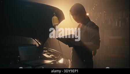 Asian Woman Technician Troubleshooting Car Electronics With Multi Screen Setup. Using Both Laptop and Tablet, Girl Monitors Engine Telemetry and Sensor Readings Simultaneously. Smart Garage of Future. Stock Photo