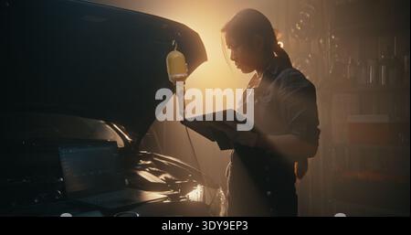 Asian Woman Technician Troubleshooting Car Electronics With Multi Screen Setup. Using Both Laptop and Tablet, Girl Monitors Engine Telemetry and Sensor Readings Simultaneously. Smart Garage of Future. Stock Photo