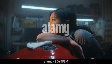 Tired But Happy Female Biker Resting Her Head on Motorcycle Tank After Repair and Polishing. Atmospheric Moment in Garage Workshop. Concept of Peace and Accomplishment in Her Private Sanctuary. Stock Photo