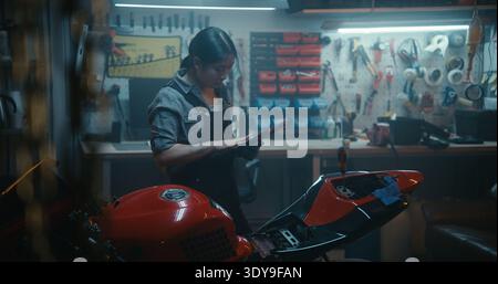 Asian Woman in Workshop Setting Uses Tablet to Follow Technical Instructions For Connecting Motorcycle Battery Cables. Professional DIY Motorcycle Repair. Concept of Technology and Mechanical Skill. Stock Photo