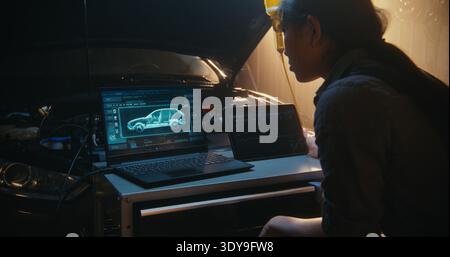 Female Technician Analyzing Vehicle Digital Twin on Laptop, With 3D Models Showing Internal Component Status. Simultaneously, Woman Interacts With Tablet to Track Performance Curves and System Logs. Stock Photo