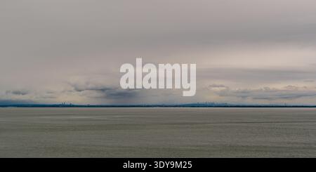 Distant view of the Vancouver skyline from the ferry, with long stretches of shoreline and clusters of tall buildings beneath a dark, heavy sky. Stock Photo