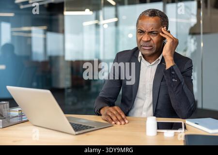 Mature businessman at modern office desk presses his temple in pain beside laptop and pill bottle, showing headache, stress and workplace overload during a tense workday Stock Photo