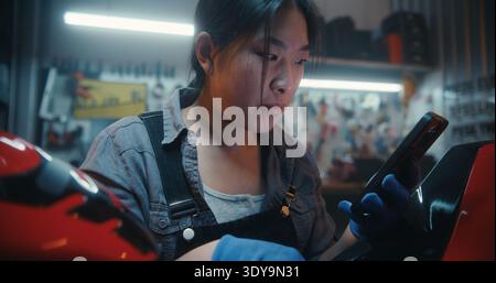 Modern Female Mechanic Integrates Technology Into Repair Process, Using AI While Assembling Motorcycle Wiring. Girl Checks Connections and Links Battery With Electronic Control System. Dolly Zoom. Stock Photo