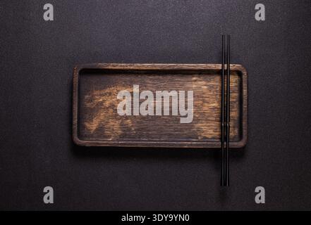 Black chopsticks are resting on a rectangular wooden board or serving tray, positioned against a dark, textured background, creating a minimalist and Stock Photo