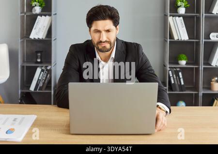 Focused businessman working on laptop at office desk, analyzing data and reviewing documents. Stock Photo