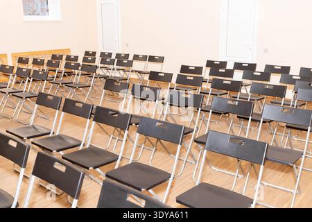 Multiple rows of identical black chairs are neatly arranged in an outdoor setting on a dirt surface, creating a uniform and symmetrical pattern. Stock Photo
