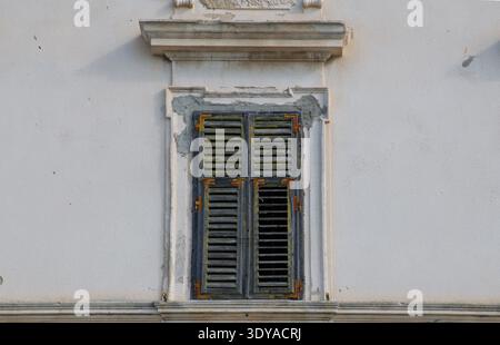 Minimalist architectural detail of a classic Mediterranean style window with white wooden shutters on a warm beige plaster wall. Soft summer sunlight. Stock Photo