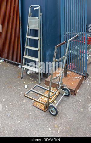 Dirty utility dolly cart and old ladder outside in city street Stock Photo