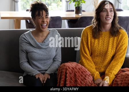 Diverse female friends laughing on gray sofa, wearing gray and mustard sweaters with rust throw Stock Photo