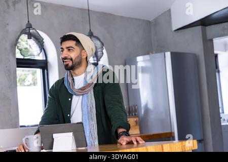 Man in late 20s standing at wooden kitchen island, wearing beanie and scarf, using tablet Stock Photo
