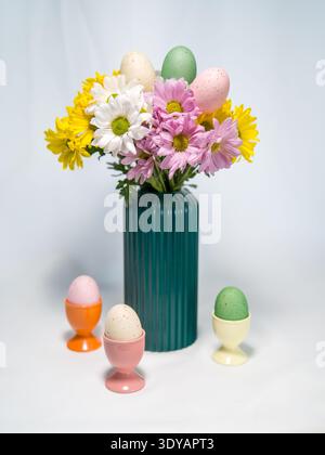Festive spring bouquet of colorful blooming daisies in a green ribbed vase with three blurred speckled Easter eggs in cups on a white table. Stock Photo