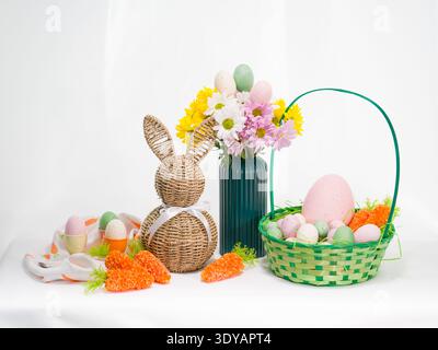 Festive Easter composition featuring a woven bunny, a basket filled with colorful eggs and carrots, and a floral bouquet on a white table. Stock Photo
